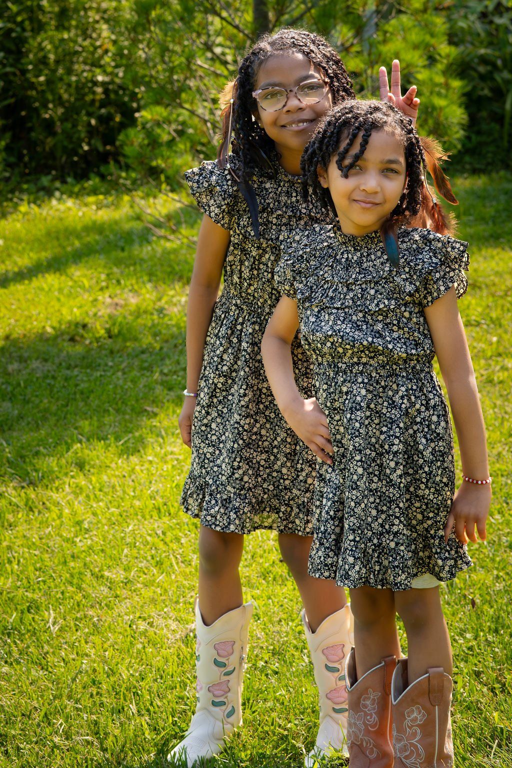 Two sisters in matching dresses and cowboy boots standing in a sunny meadow